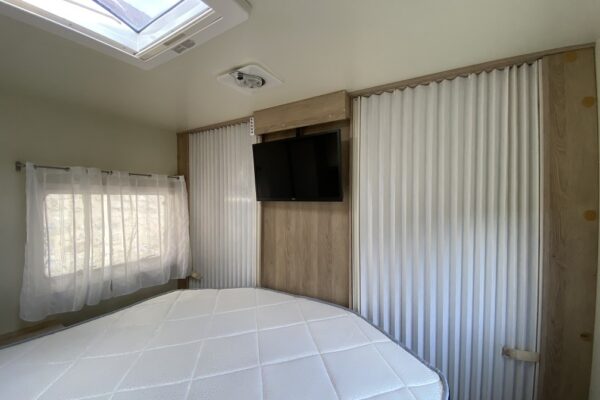 Helles Zimmer mit Bett, Fenster, Fernseher und weißen Vorhängen.