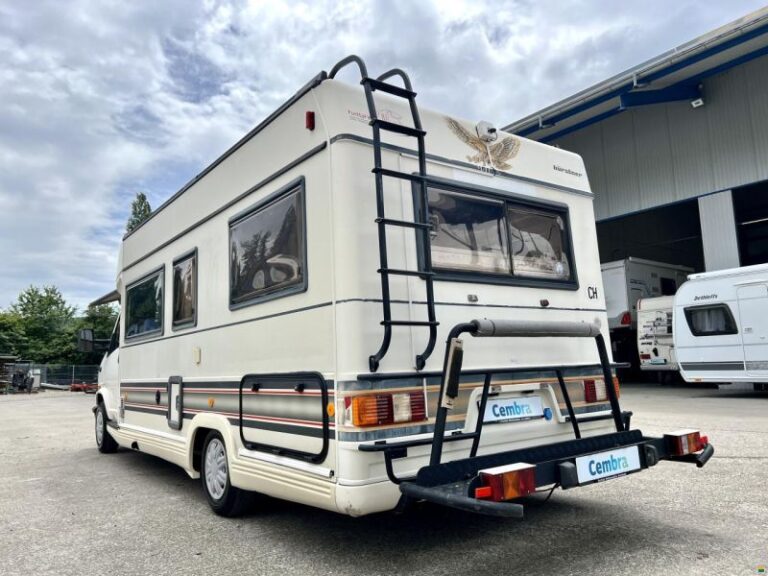 Camping-car blanc stationné, échelle arrière, ciel partiellement nuageux.