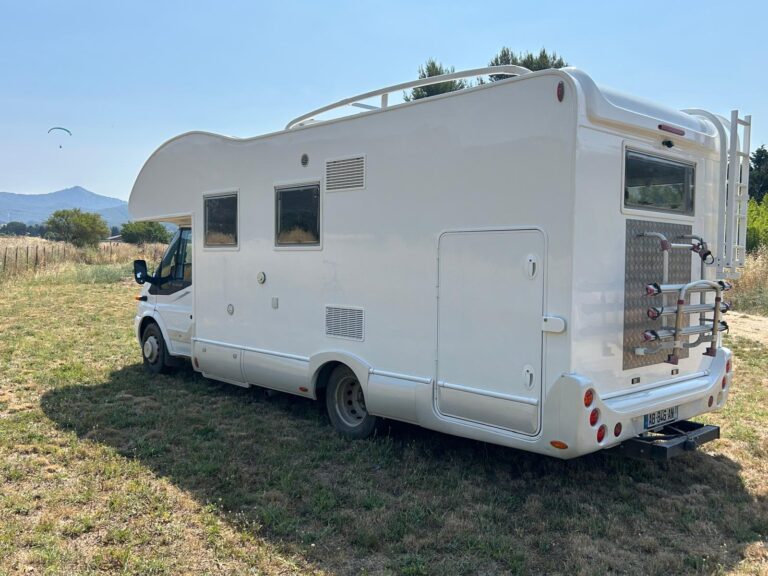 Camping-car blanc sur terrain herbeux avec montagnes en arrière-plan.