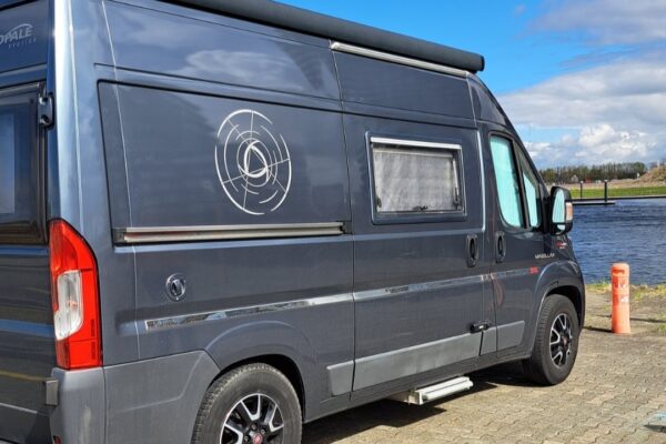 Furgone camperizzato parcheggiato vicino a un fiume sotto un cielo azzurro.