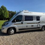 Camping-car argenté sur gravier, arbres en arrière-plan.