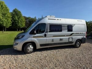 Camping-car argenté sur gravier, arbres en arrière-plan.