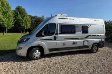 Camping-car argenté sur gravier, arbres en arrière-plan.