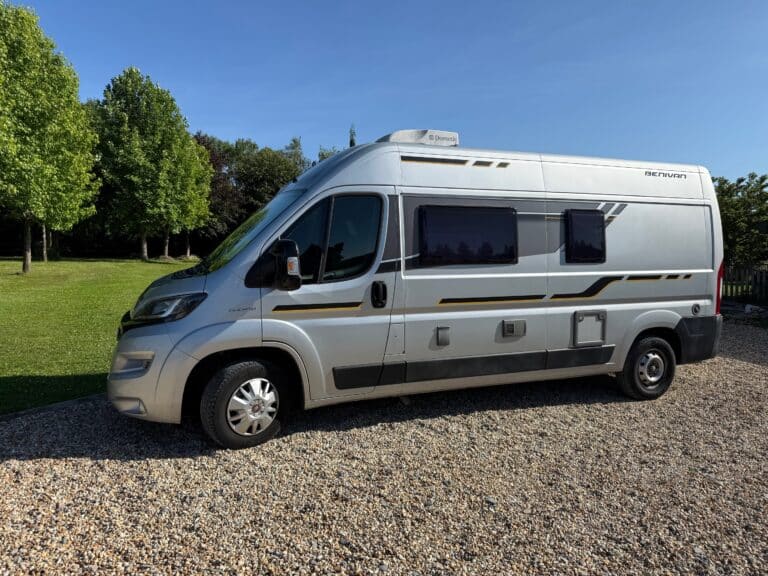 Camping-car argenté sur gravier, arbres en arrière-plan.
